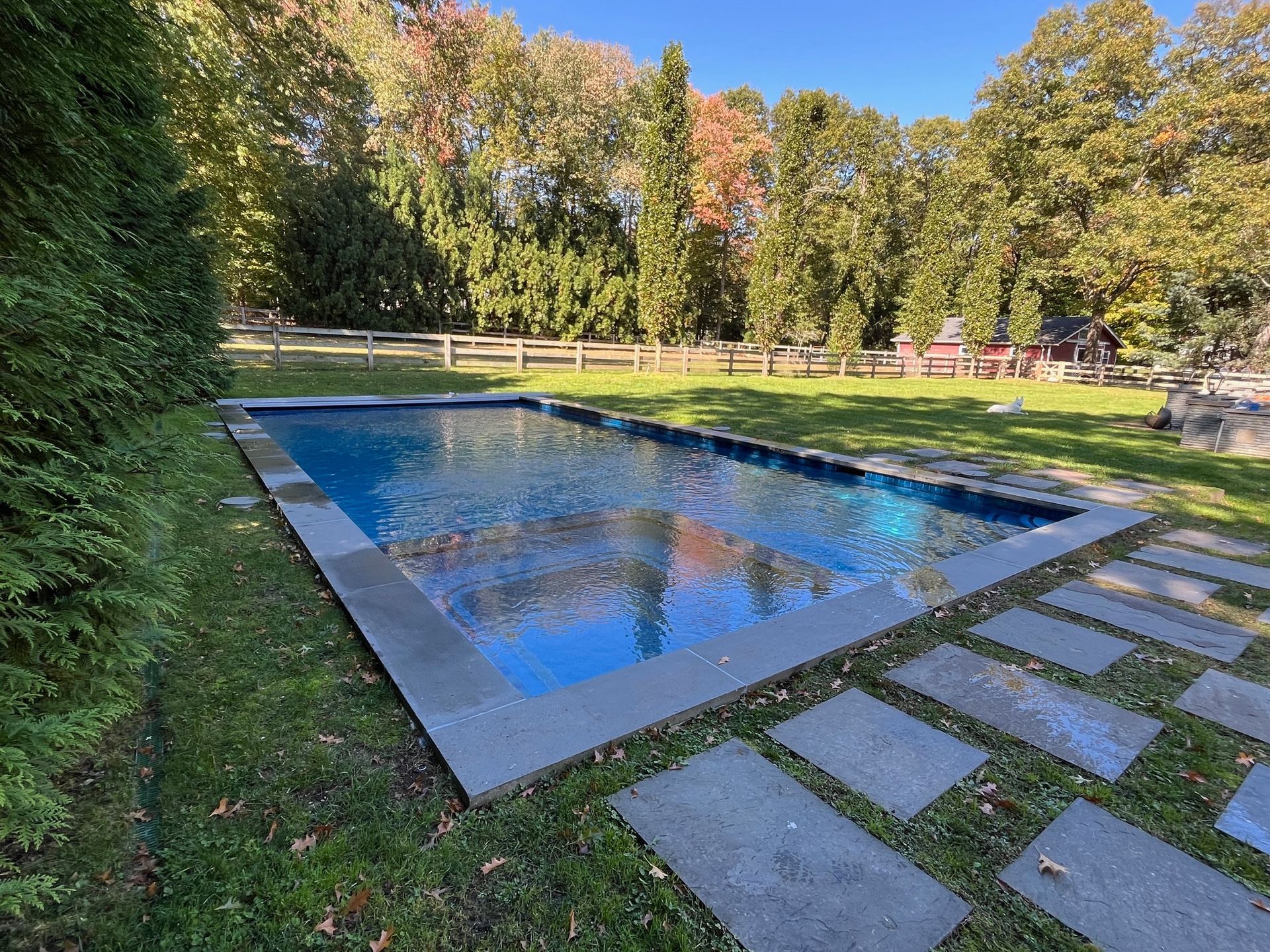 A rectangular swimming pool surrounded by a green lawn and autumn trees, with grey stone stepping stones in the foreground.