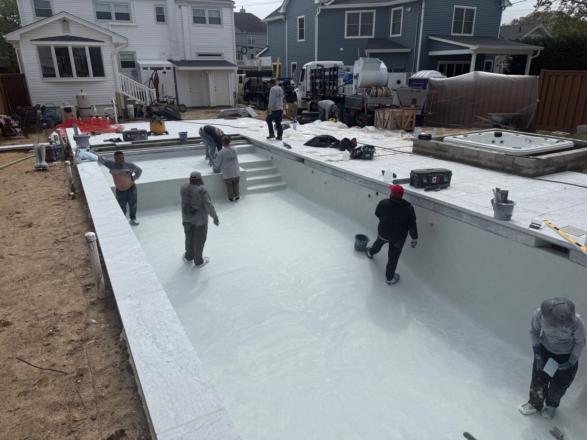 Workers in a residential backyard applying a white finish to the interior of an in-ground swimming pool.