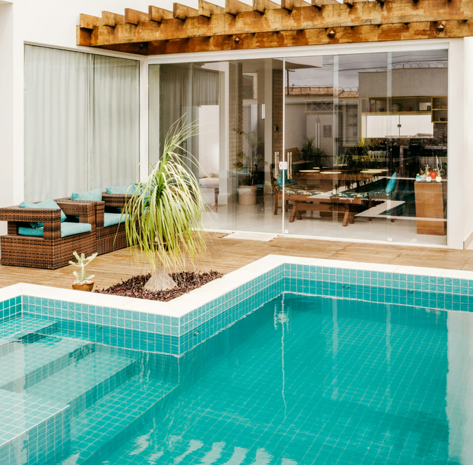 A turquoise tiled pool with a small potted palm tree on the deck outside a glass-walled home with a wooden pergola.