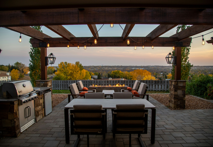 A patio at sunset with a dining table, outdoor seating, a built-in grill, and a fire pit overlooking a scenic view.