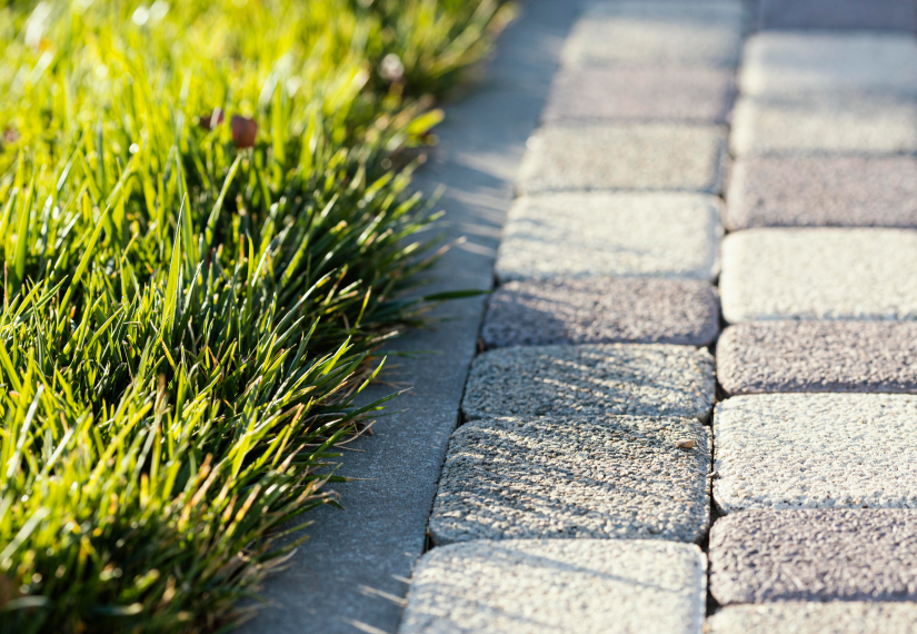 A close-up view of a green grass lawn meeting a paved walkway made of textured, light-colored stone pavers.