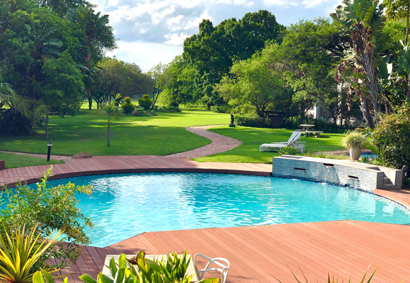 A vibrant blue swimming pool with a reddish deck, surrounded by a lush green lawn and trees under a sunny sky.