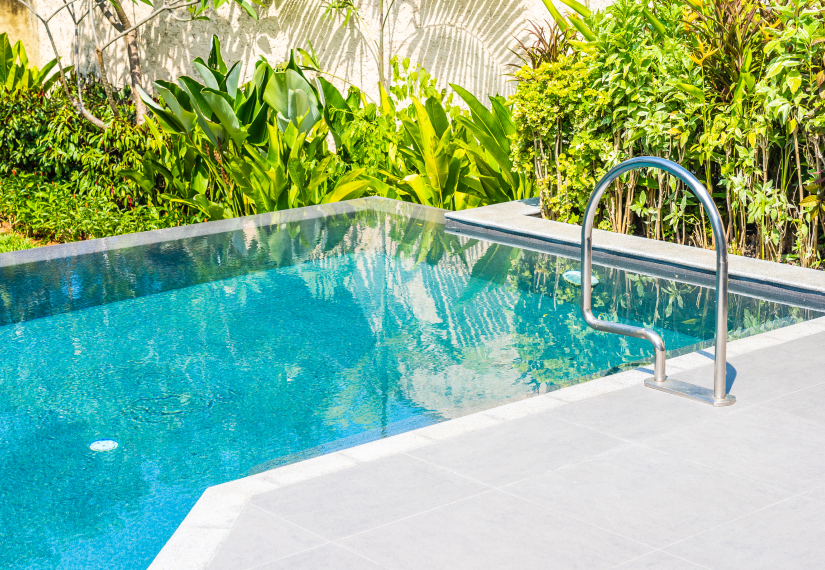 A rectangular swimming pool with bright blue water and a silver handrail, surrounded by lush green foliage.