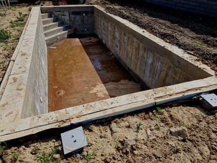 A rectangular, unfinished concrete swimming pool in a dirt construction site with concrete stairs leading inside.