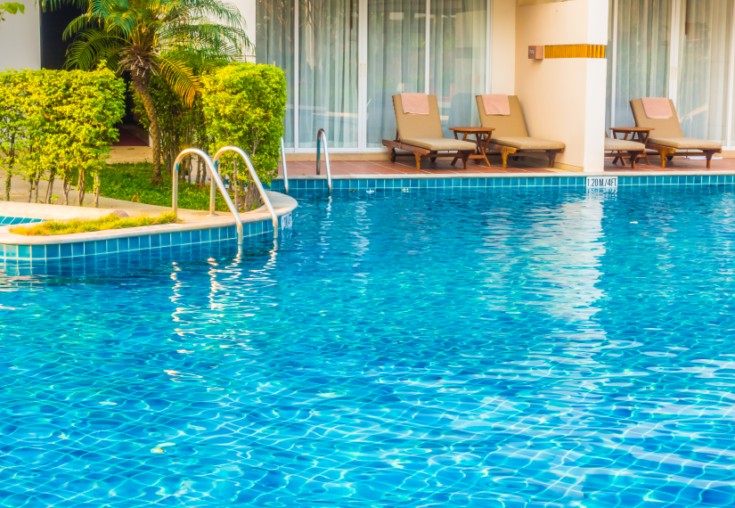 A resort swimming pool with a metal ladder, surrounding lush green landscaping, and lounge chairs on the patio.