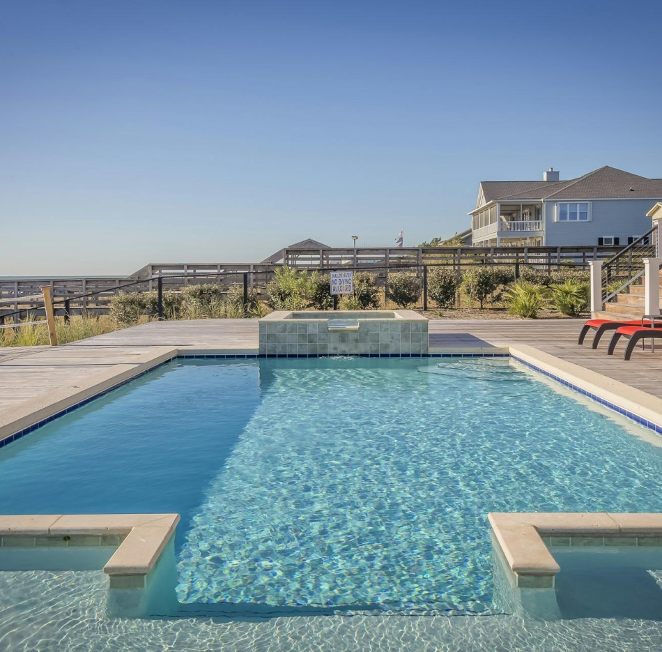 A rectangular swimming pool with a stone hot tub on a sunny deck, featuring blue water and a residential home in the back.