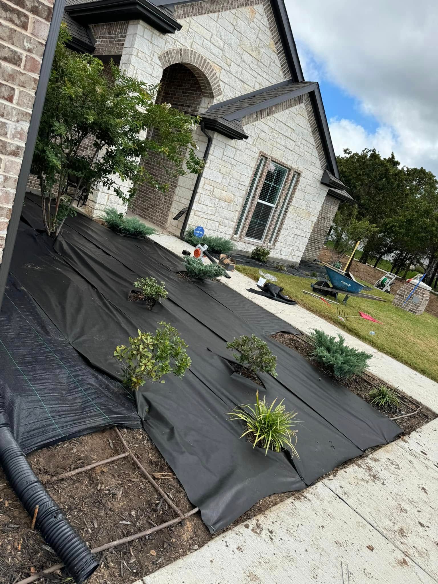 Front yard with black landscape fabric, small shrubs, and a stone house under a cloudy sky
