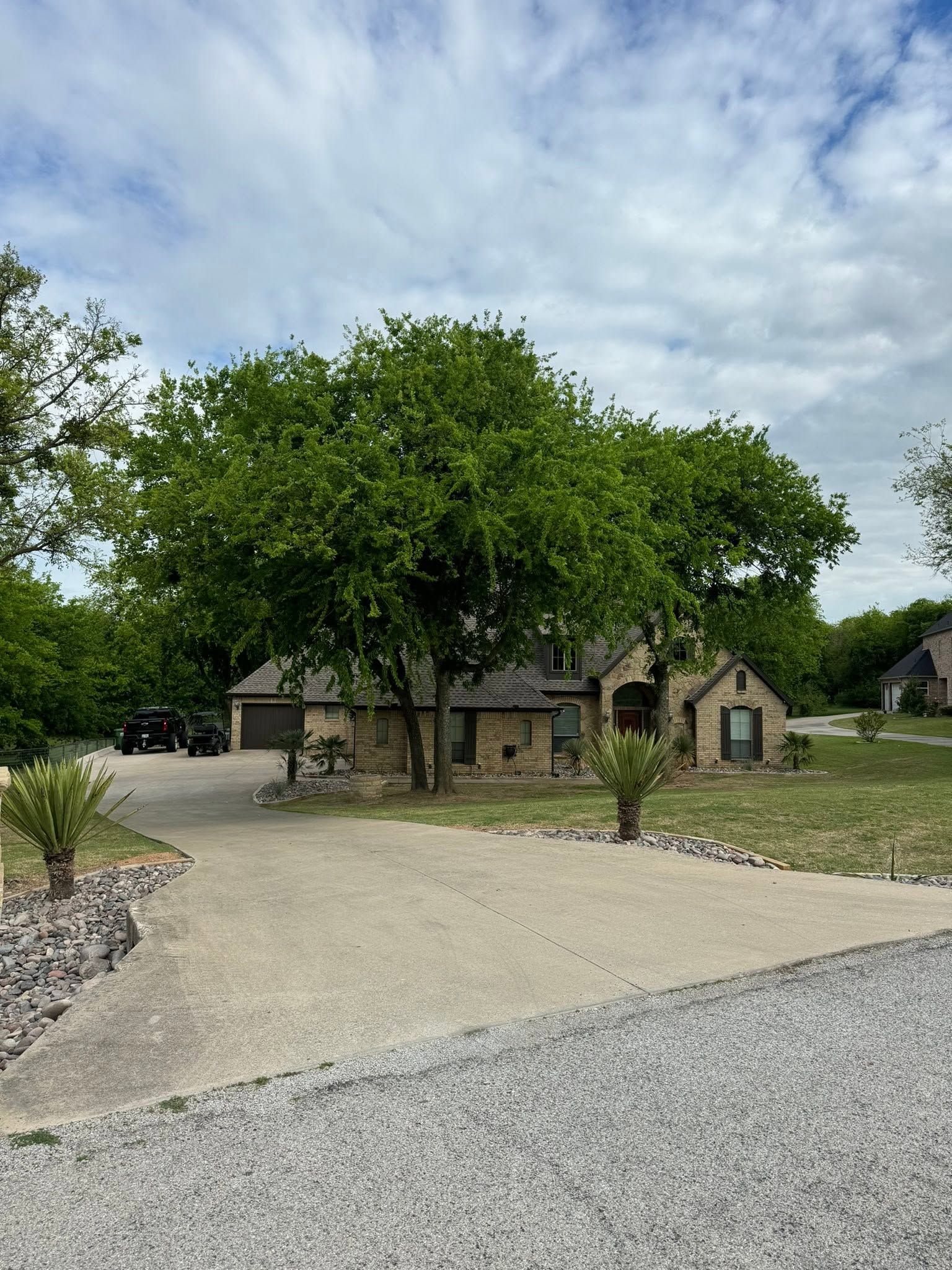 Stone cottage with a large tree, gravel driveway, and cloudy sky in a rural setting