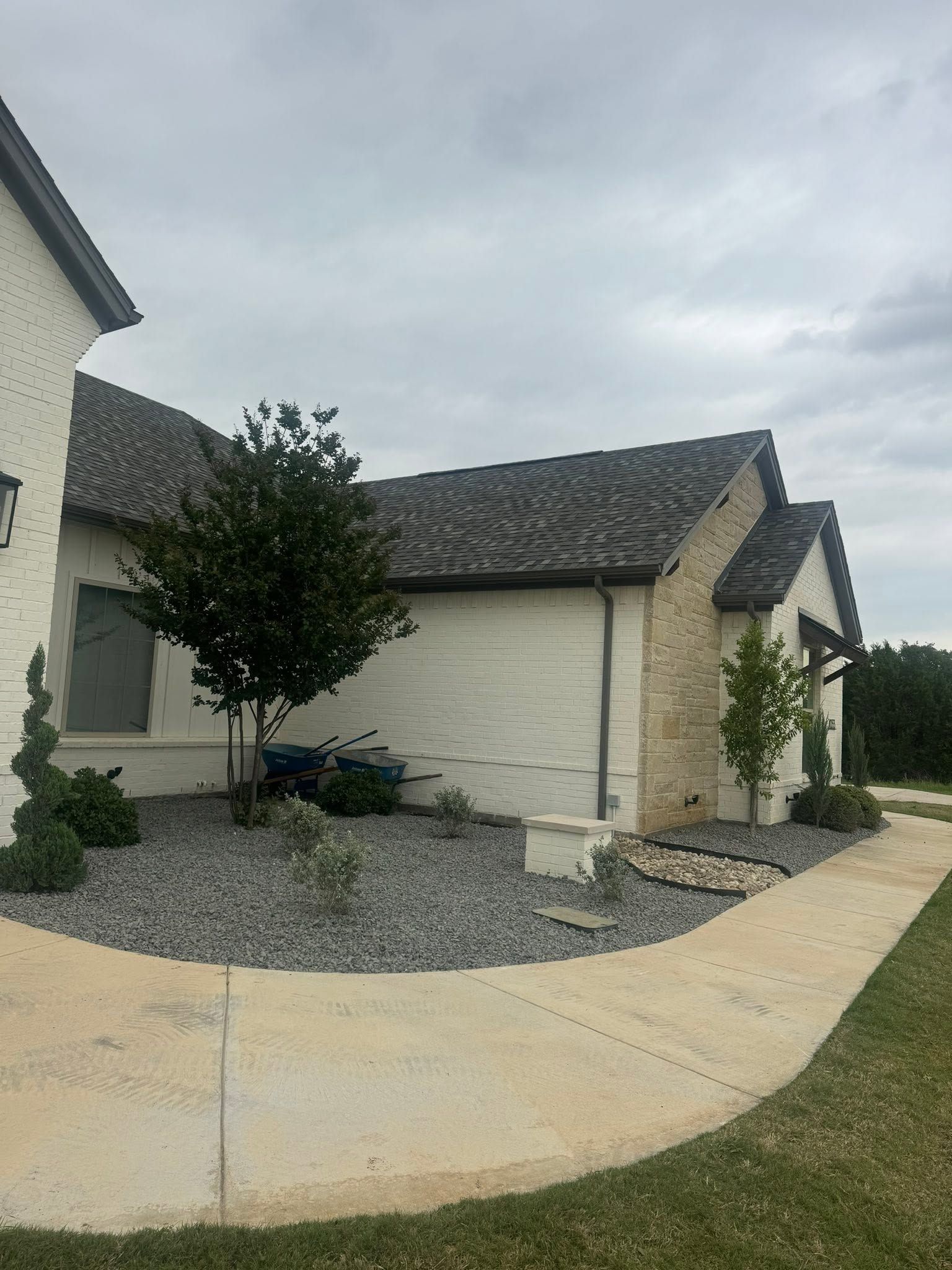 Suburban house exterior with light stucco walls, dark roof, and a small front yard with gravel and a tree