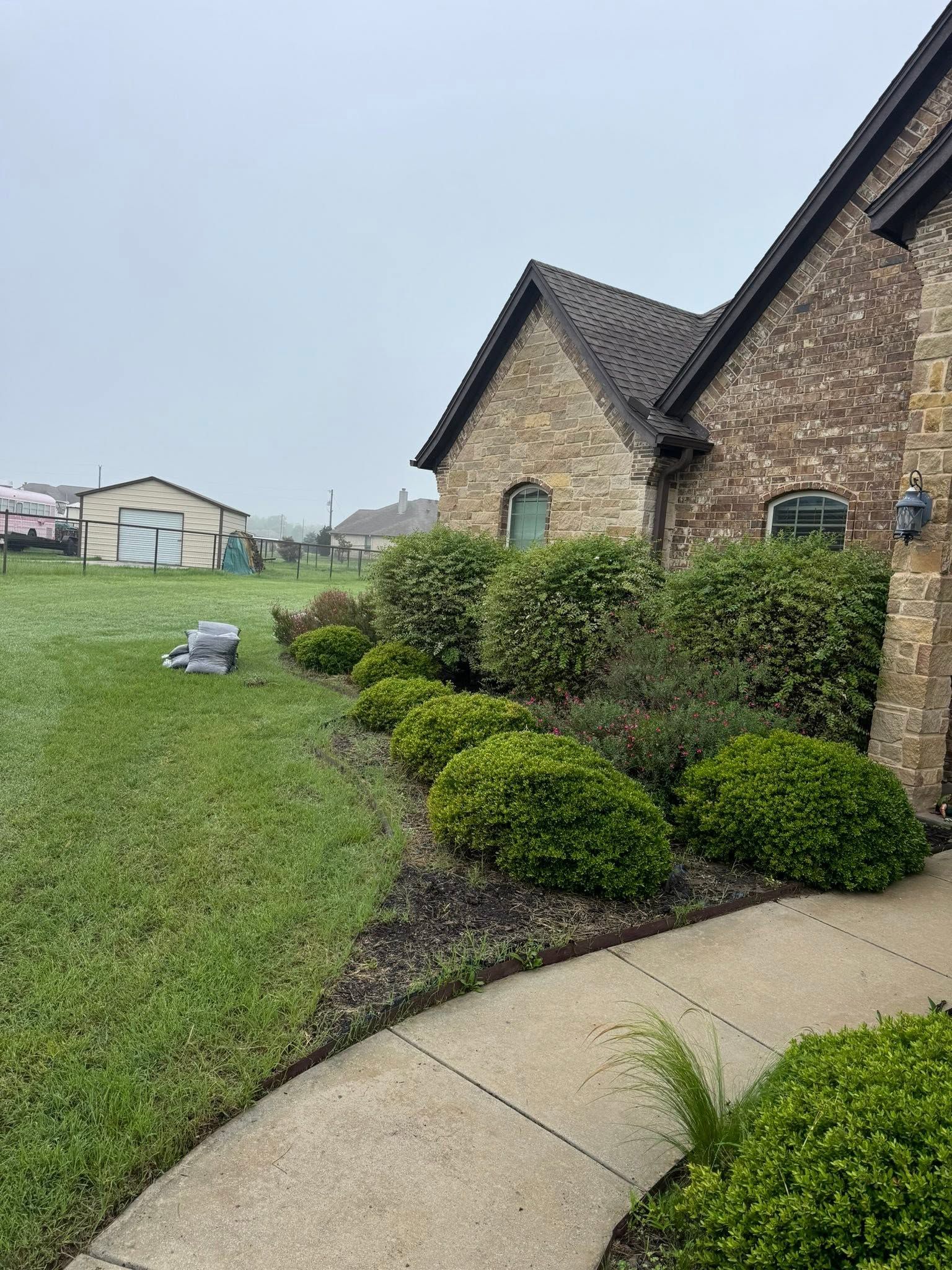 Brick houses beside a curved sidewalk and trimmed shrubs along a grassy yard under a cloudy sky