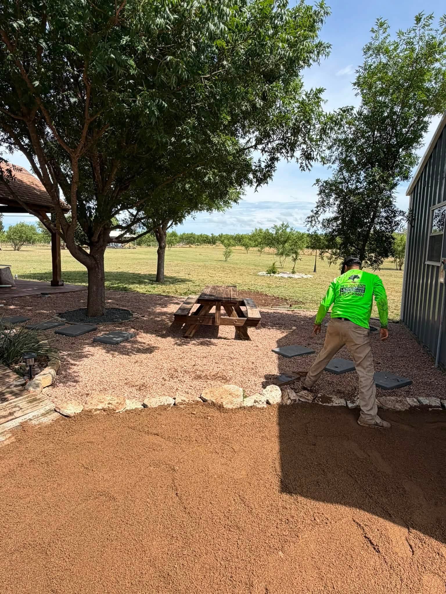 Outdoor yard with a tree, picnic table, and a worker in a neon green shirt near a gravel path.