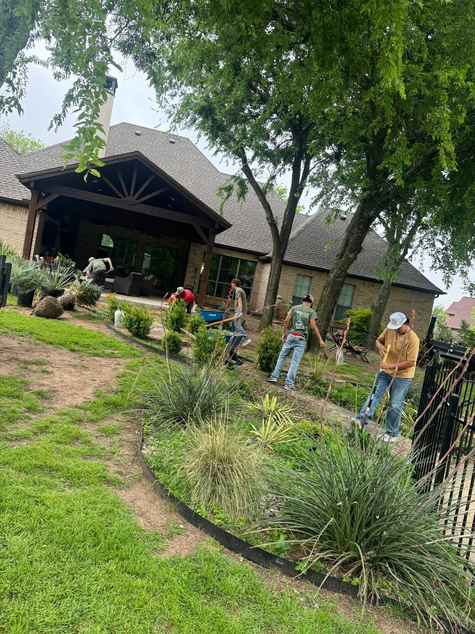 People gardening in a lush yard beside a rustic house with trees and flower beds