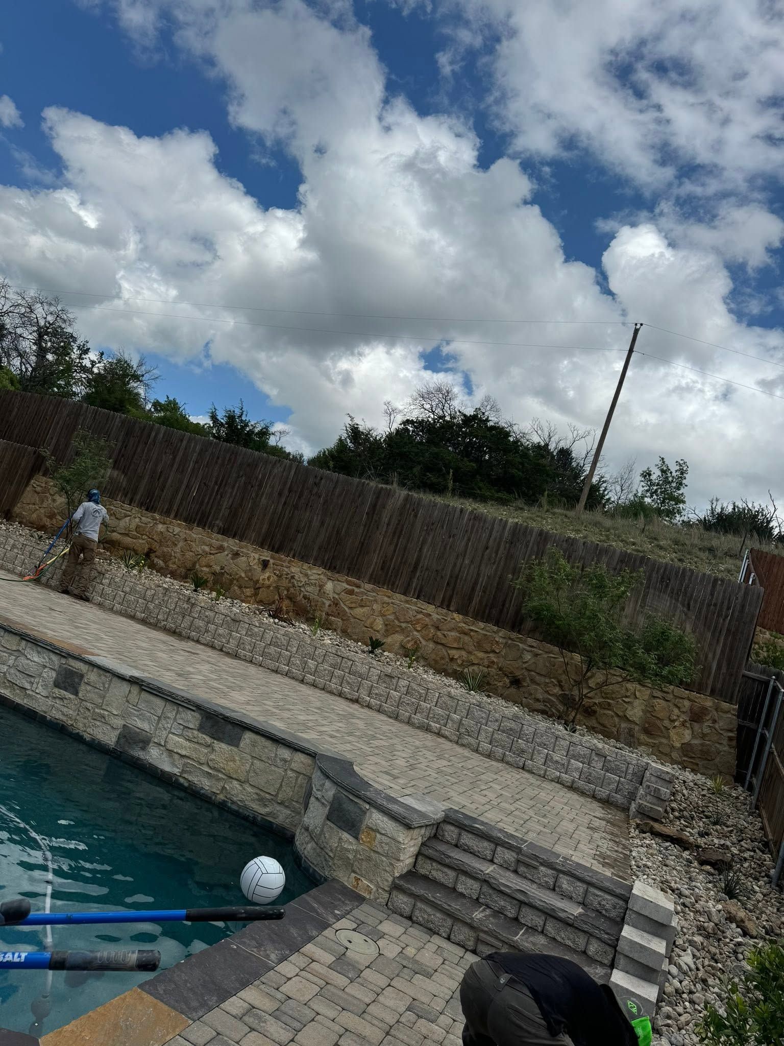 Backyard swimming pool beside a stone wall under a cloudy sky, with a person bent over near the water.