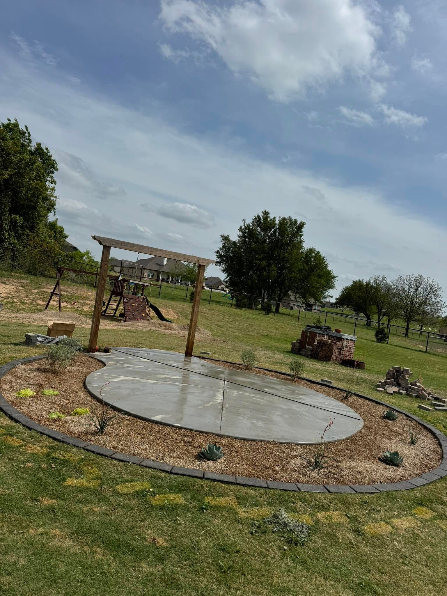 Small backyard patio with a round concrete pad, pergola frame, and landscaped garden under a cloudy sky