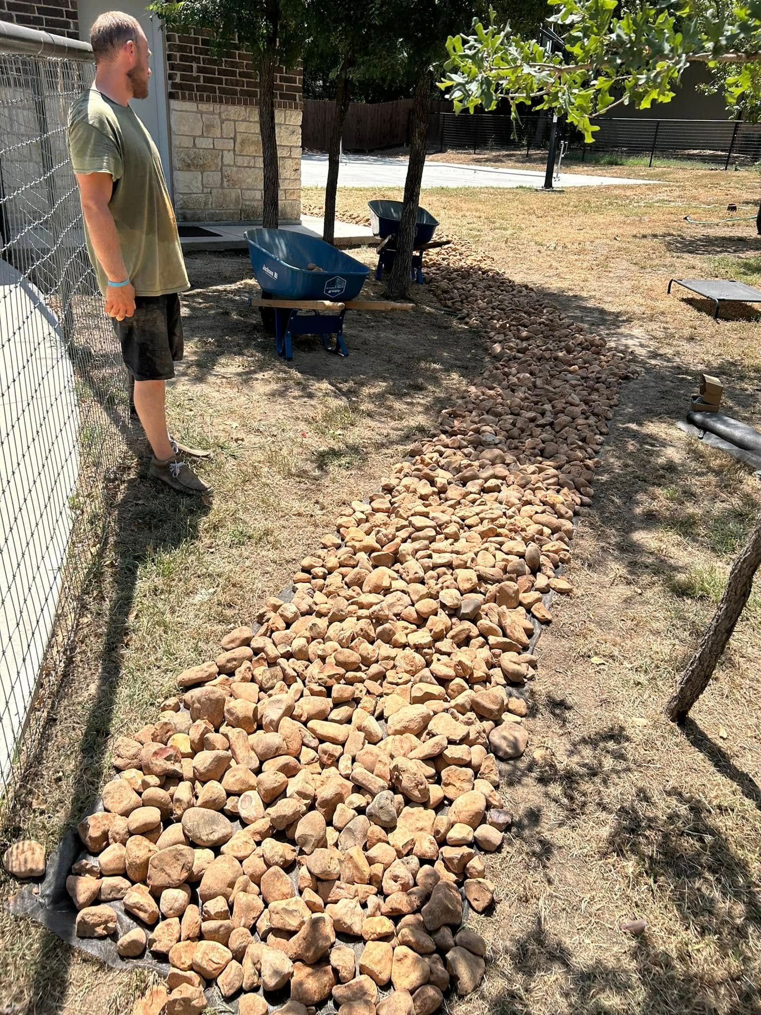 Man standing beside a wheelbarrow and a rock-lined path in a dry yard under trees