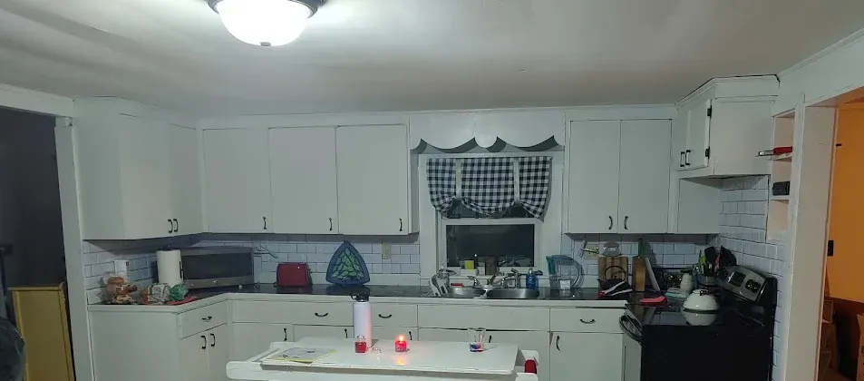 A white kitchen with overhead cabinets, a stove, and a window with a checkered curtain.