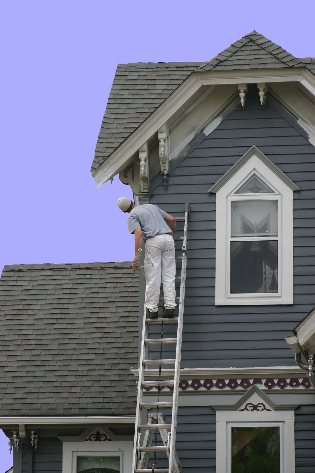 Man on ladder painting the side of a blue Victorian house.