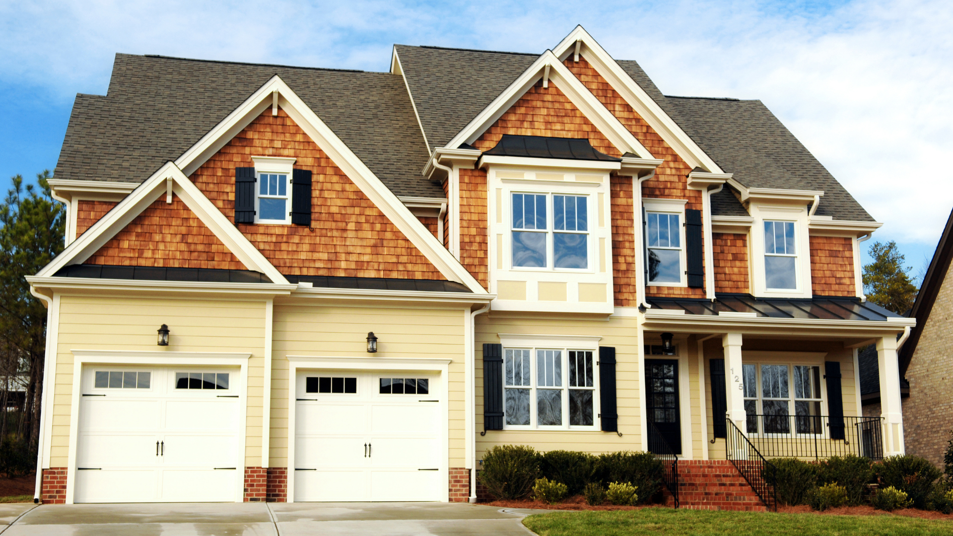 Two-story house with tan siding, brown wooden accents, two-car garage, and black shutters.
