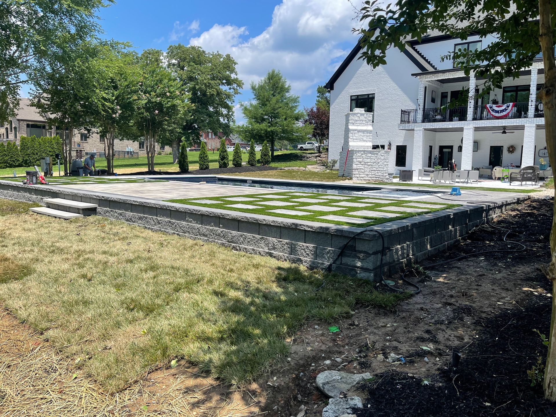 Stone raised patio in a grassy yard beside a white building and trees under a sunny sky.