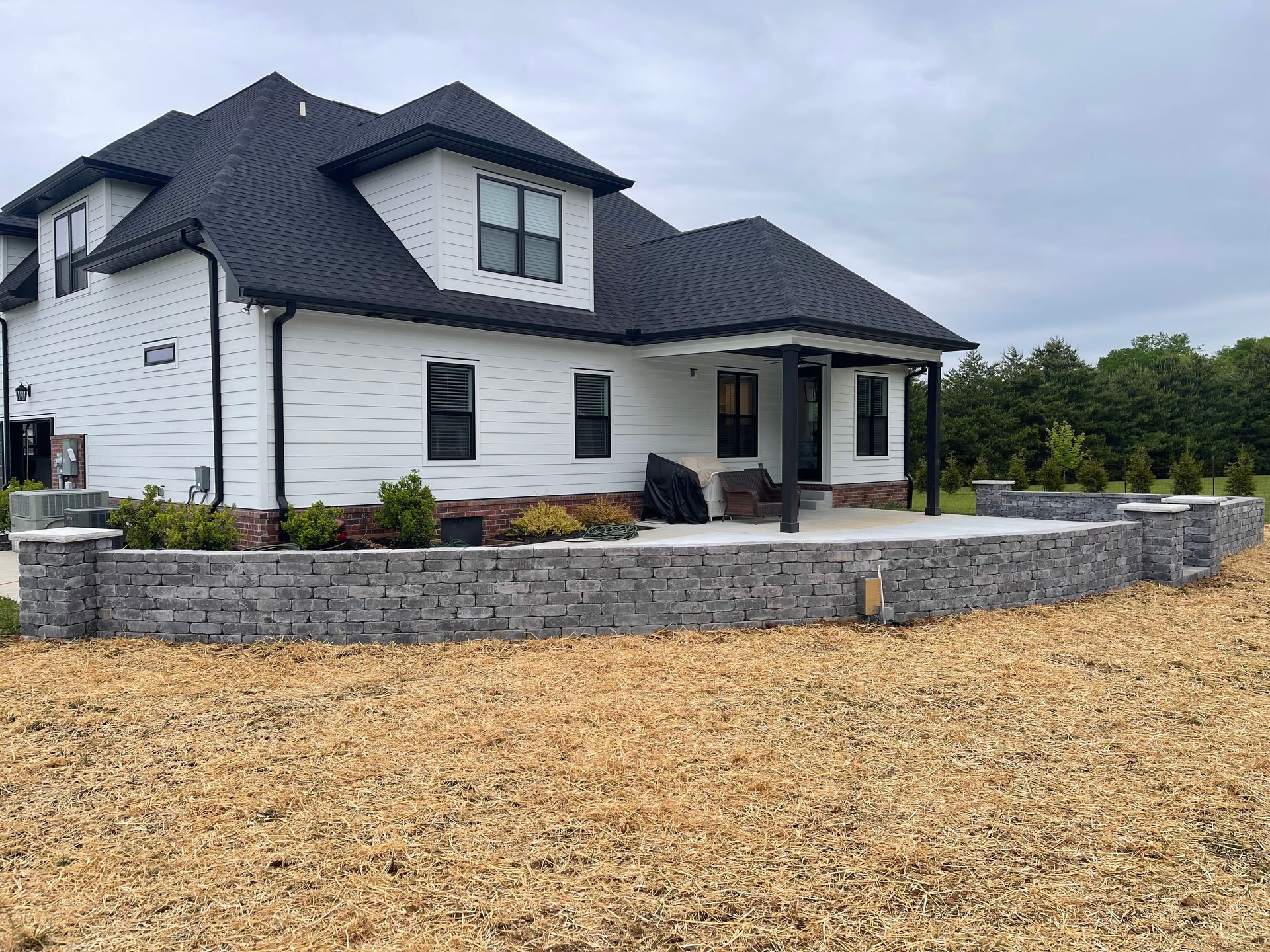White modern house with dark roof, stone retaining wall, and gravel yard under overcast sky