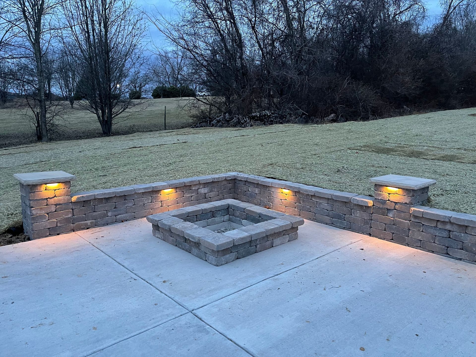 Stone patio with built-in fire pit and lit wall sconces overlooking a grassy yard at dusk