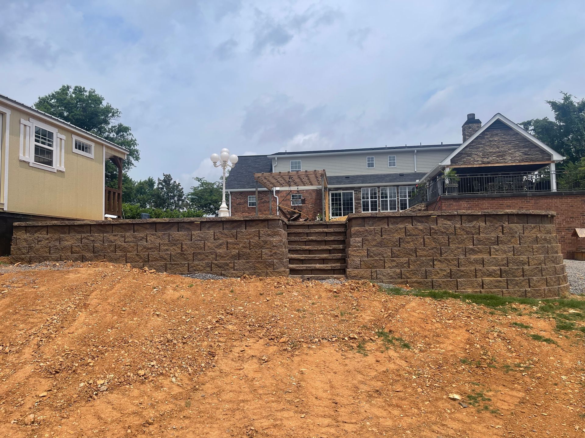 Two houses behind a brick retaining wall and steps, with a dirt foreground and cloudy sky.