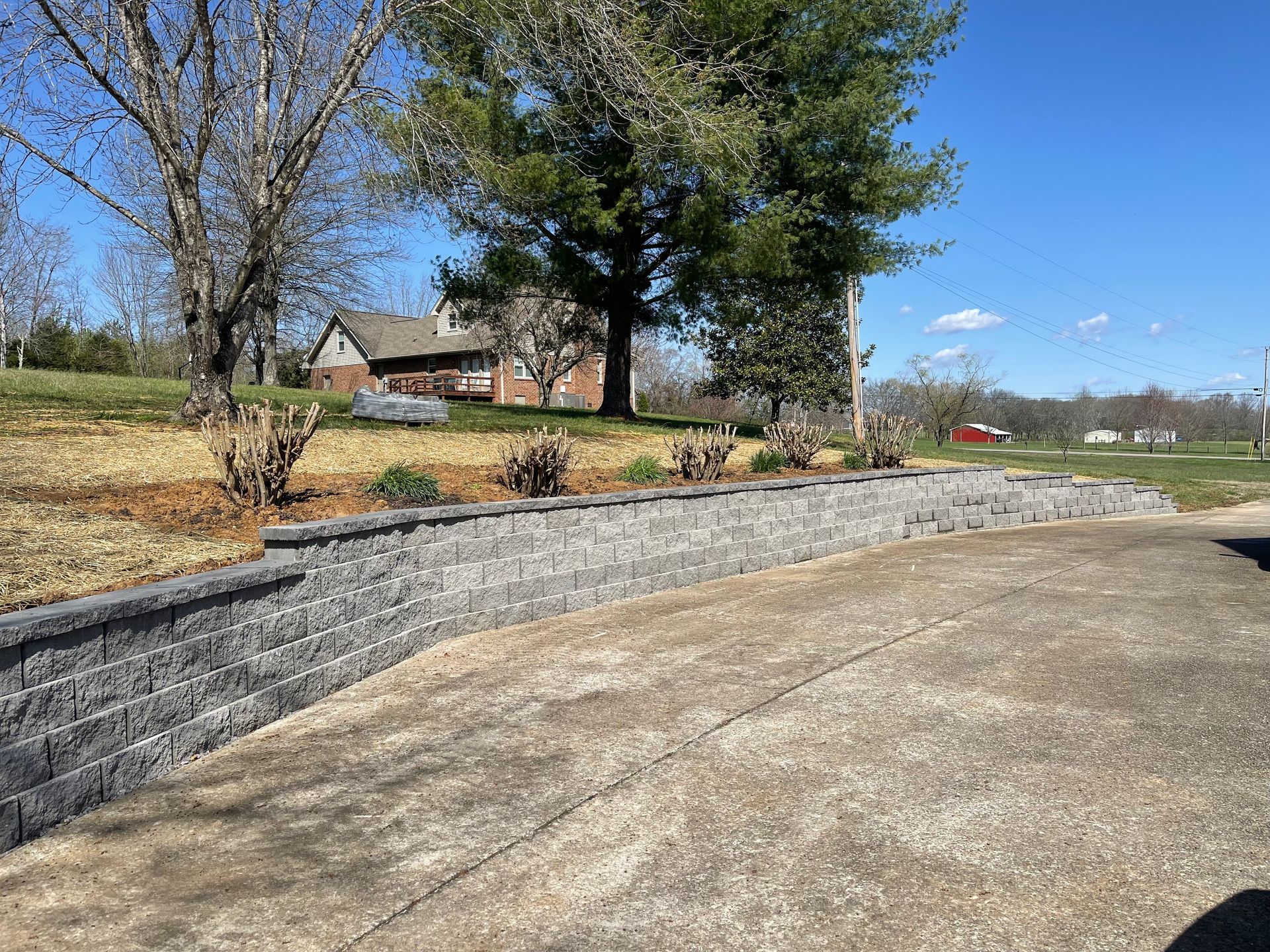 Gravel drive beside a stone retaining wall, with a house, trees, and blue sky in the background