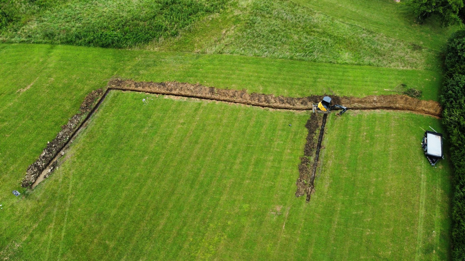 Aerial view of a grassy field with an L-shaped stone wall and a small tractor near the top right.