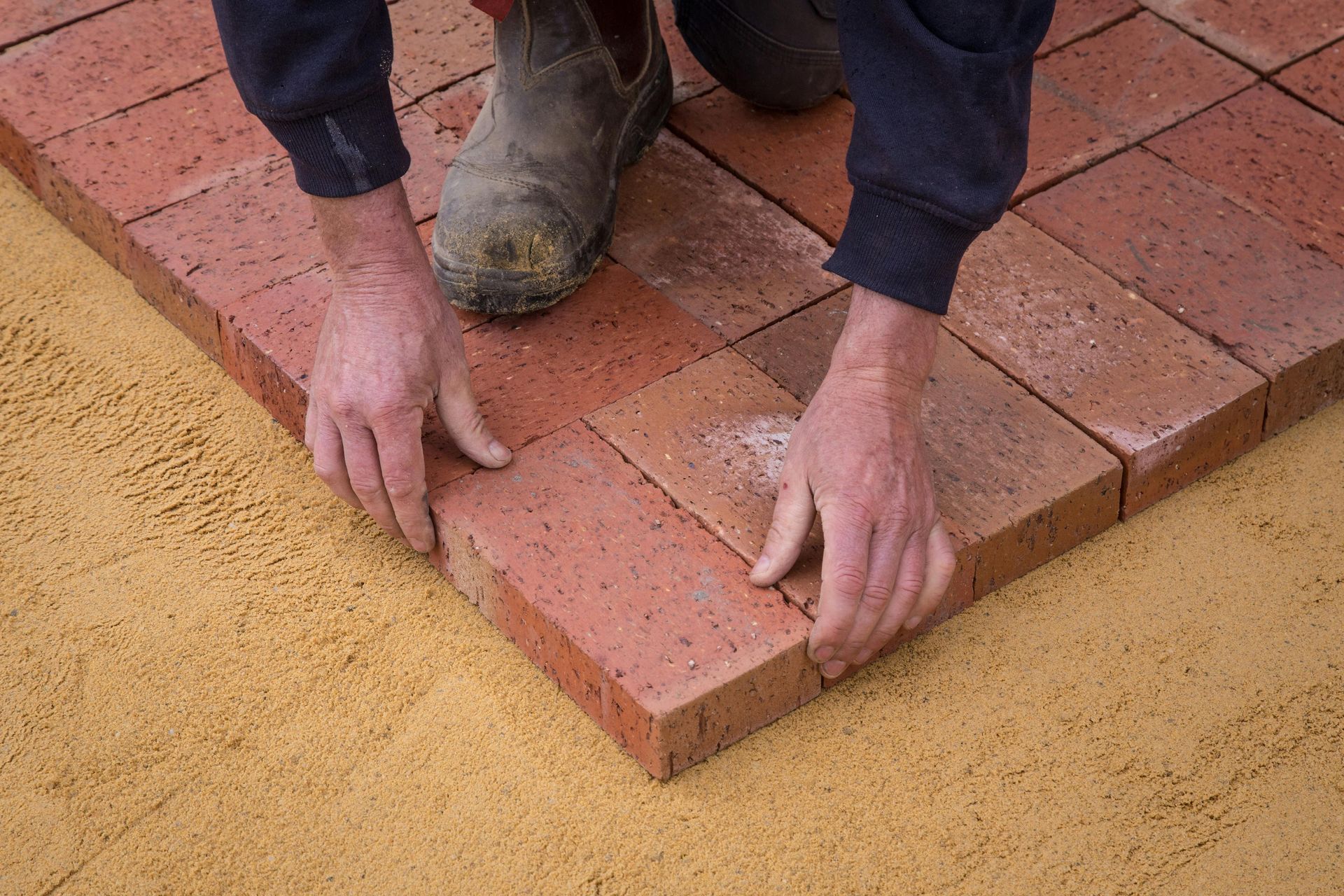 Person laying red bricks on sand with both hands and a boot visible nearby