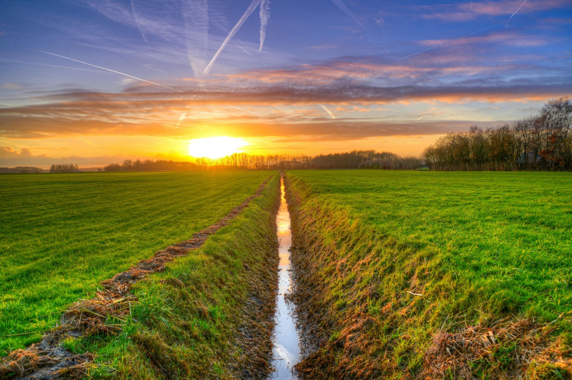 Sunset over green fields with a narrow drainage ditch reflecting the sky