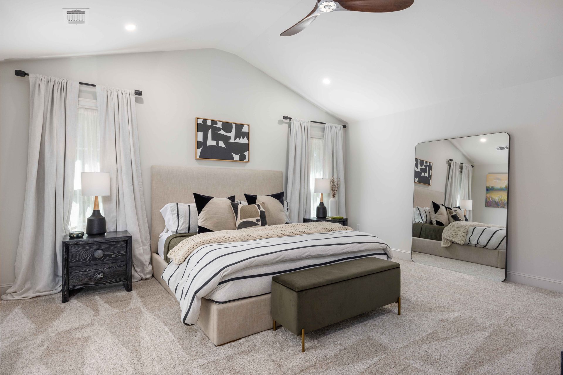 Bright bedroom with a tufted bed, bench, mirrored dresser, and soft white curtains on carpeted floor