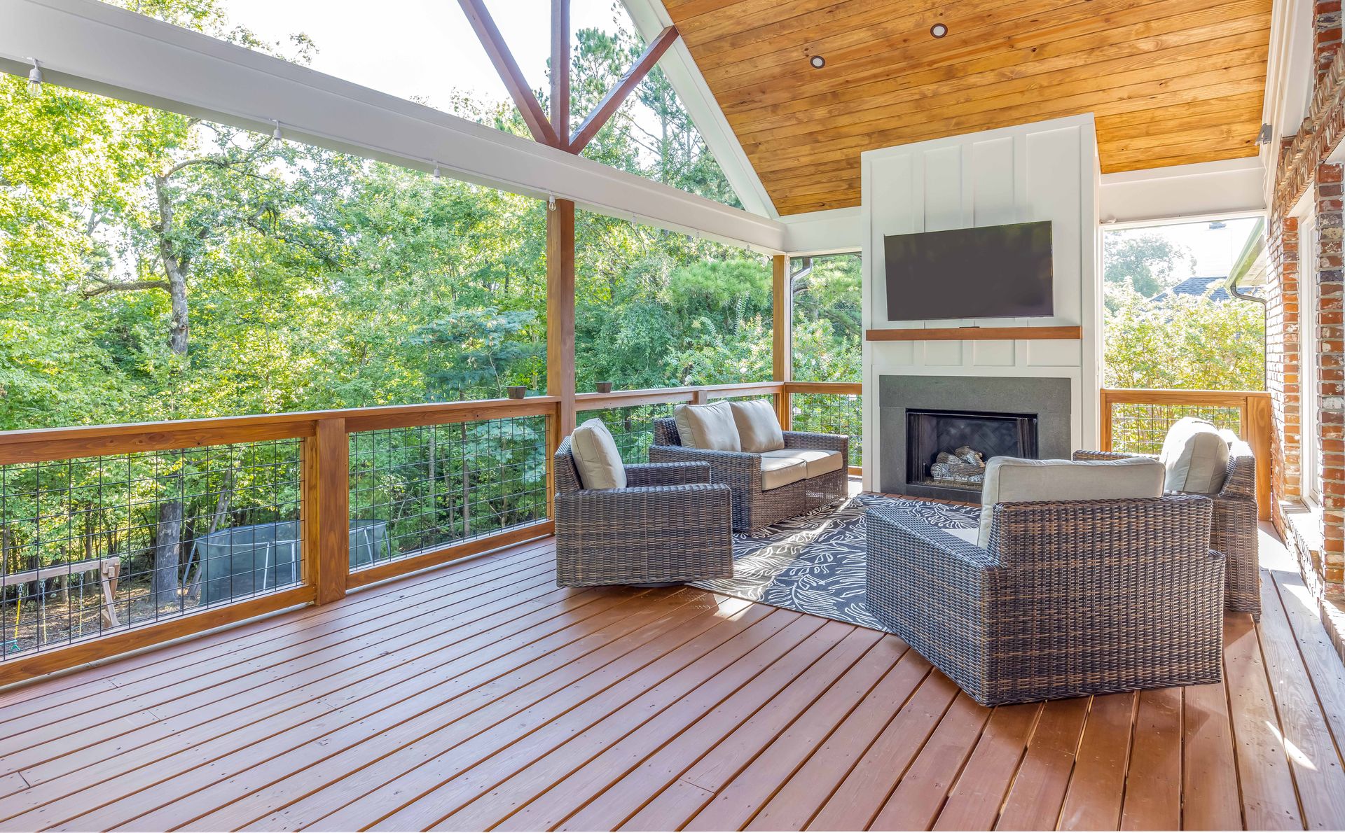 Covered wooden deck with wicker chairs and fireplace, overlooking green trees through open screened walls