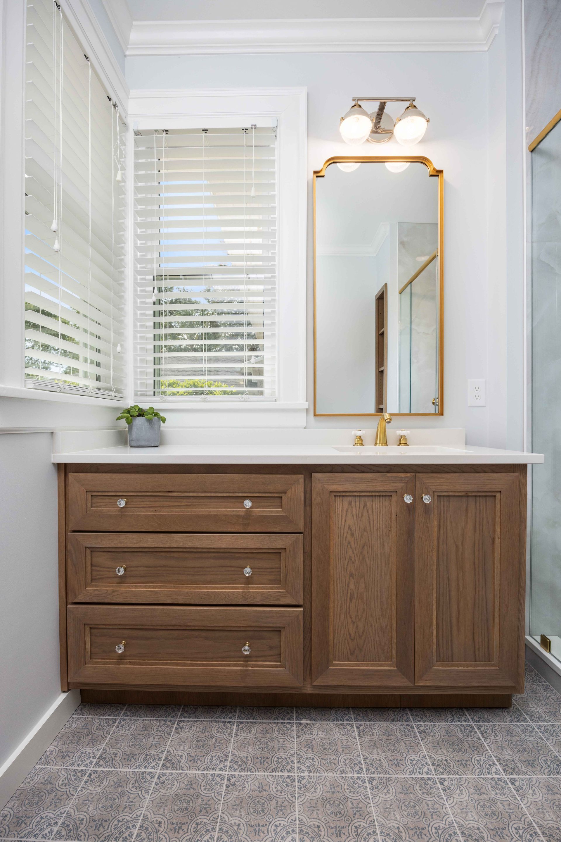 Bathroom vanity with wood cabinets, white countertop, gold-framed mirror, and window blinds