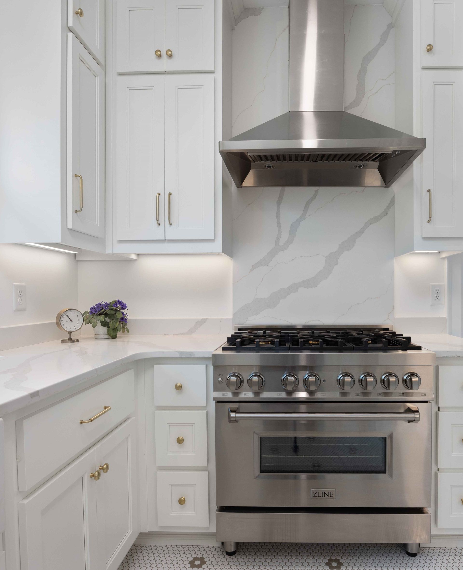 Modern kitchen with stainless steel stove and range hood, white cabinets, marble backsplash, and a small flower arrangement.