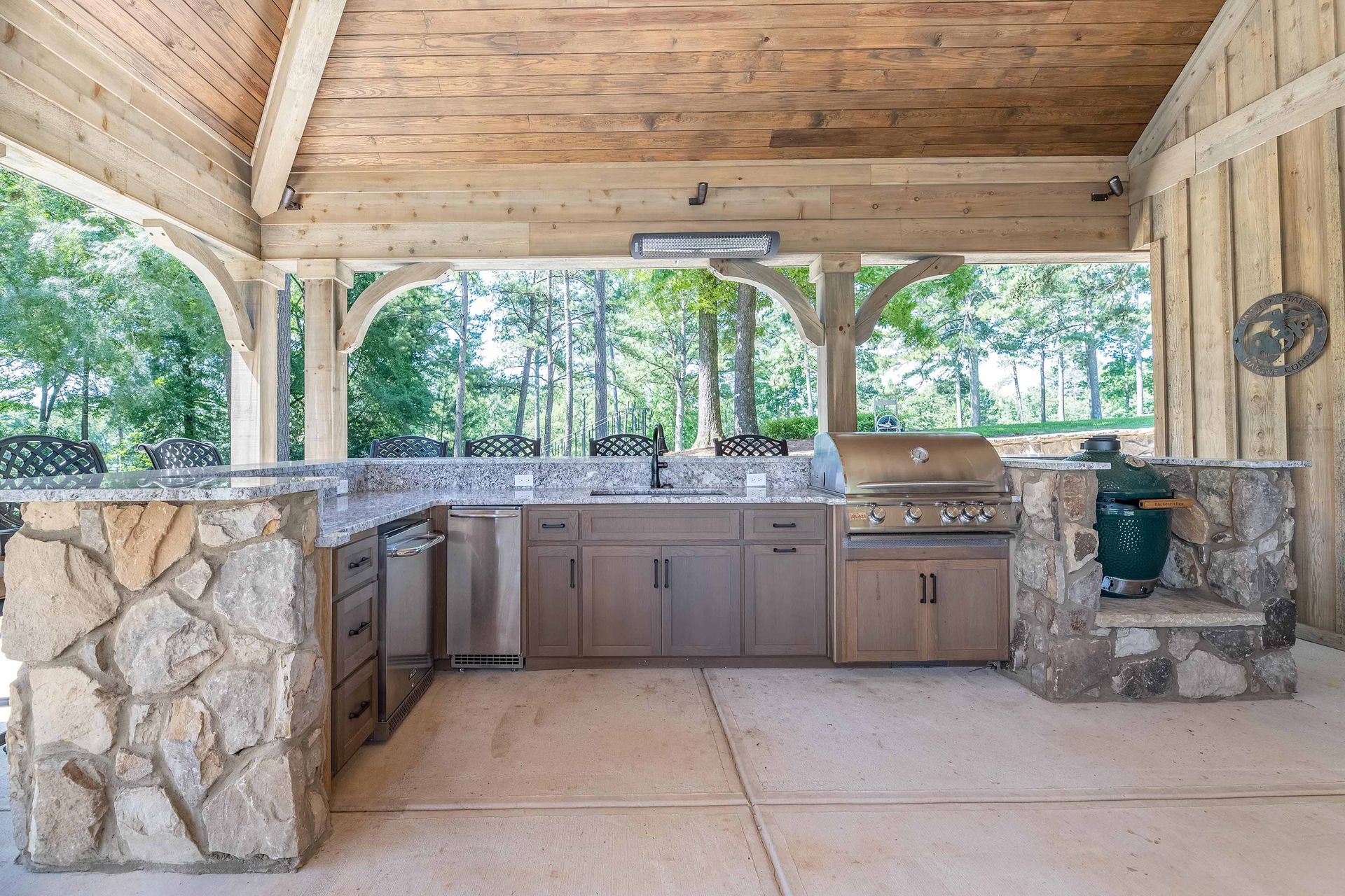 Outdoor stone kitchen with wood ceiling, beige cabinets, sink, and stone counter under a covered patio