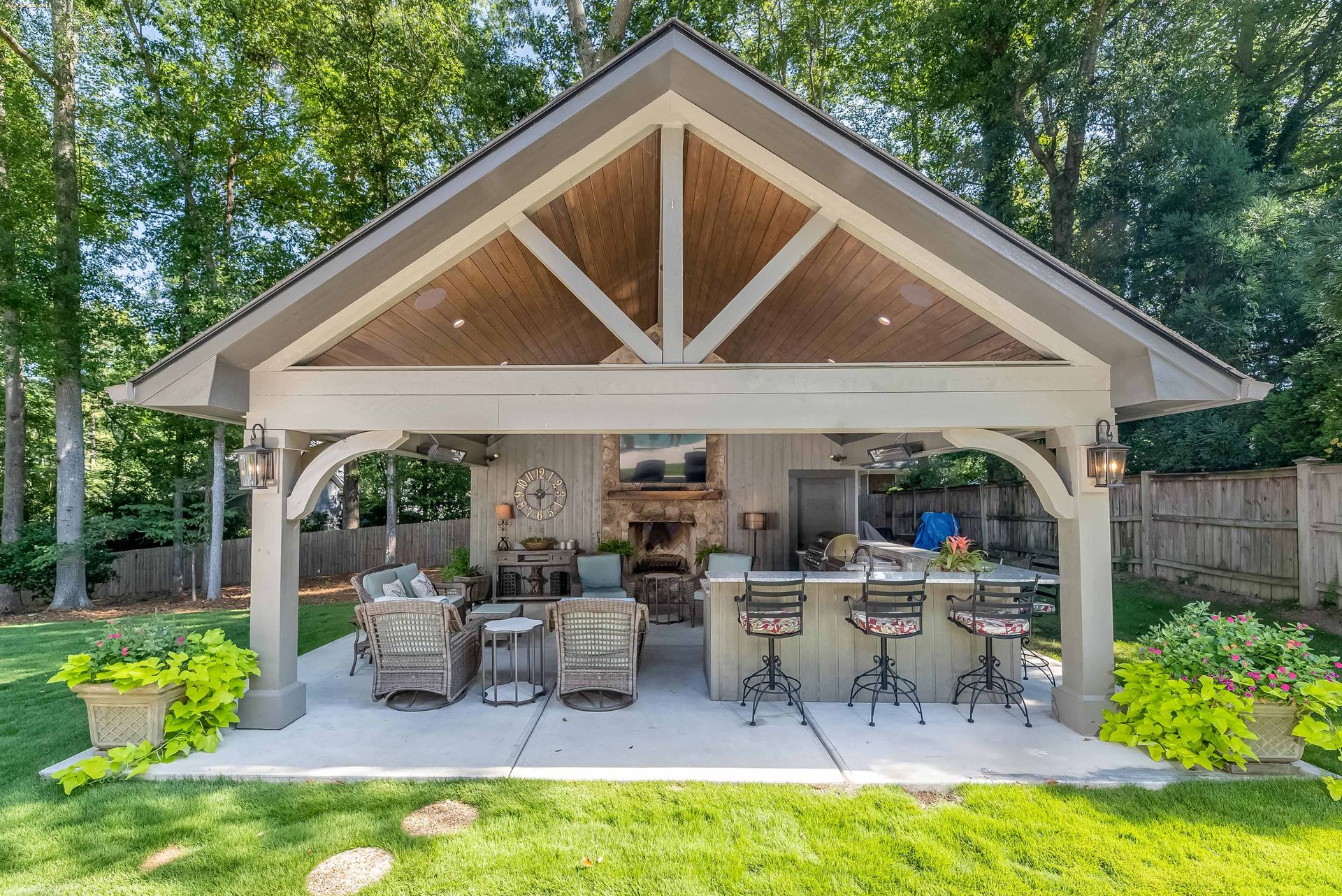 Covered outdoor patio with wicker seating, bar stools, and a stone fireplace surrounded by greenery.