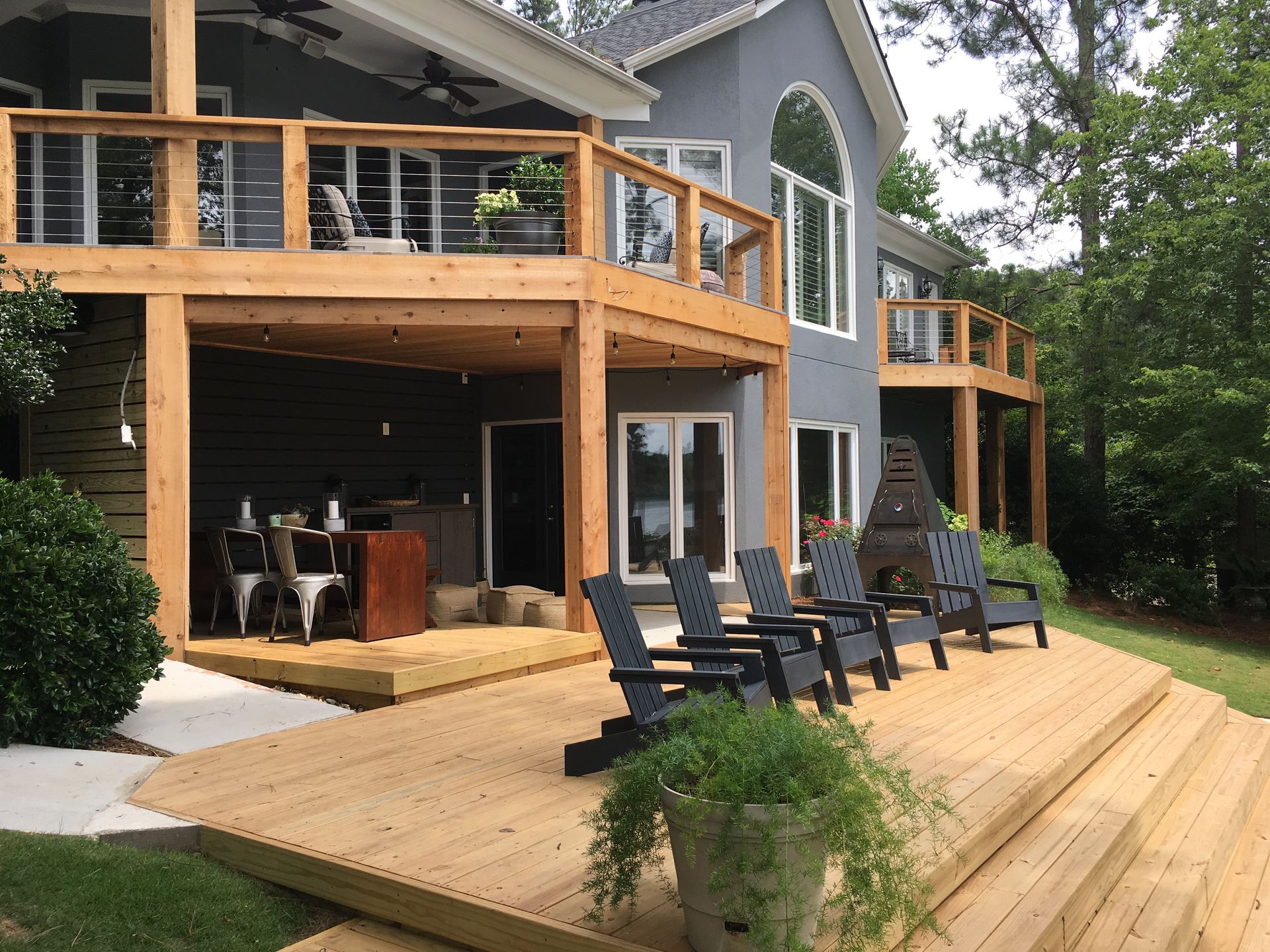 Wooden deck and patio with black chairs on a gray house, overlooking a wooded yard