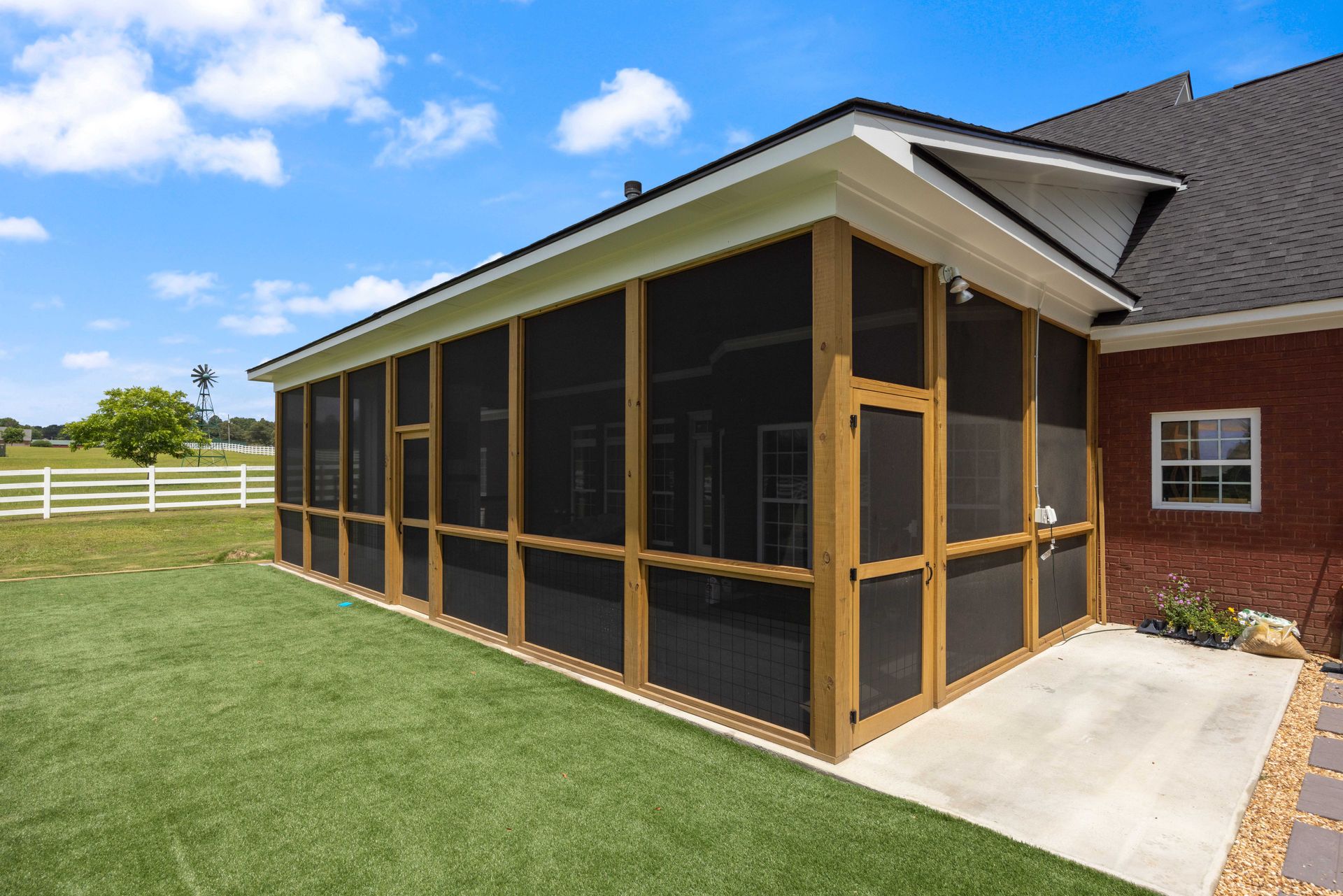 Screened porch addition with black mesh panels and wood trim beside a brick house on a sunny lawn