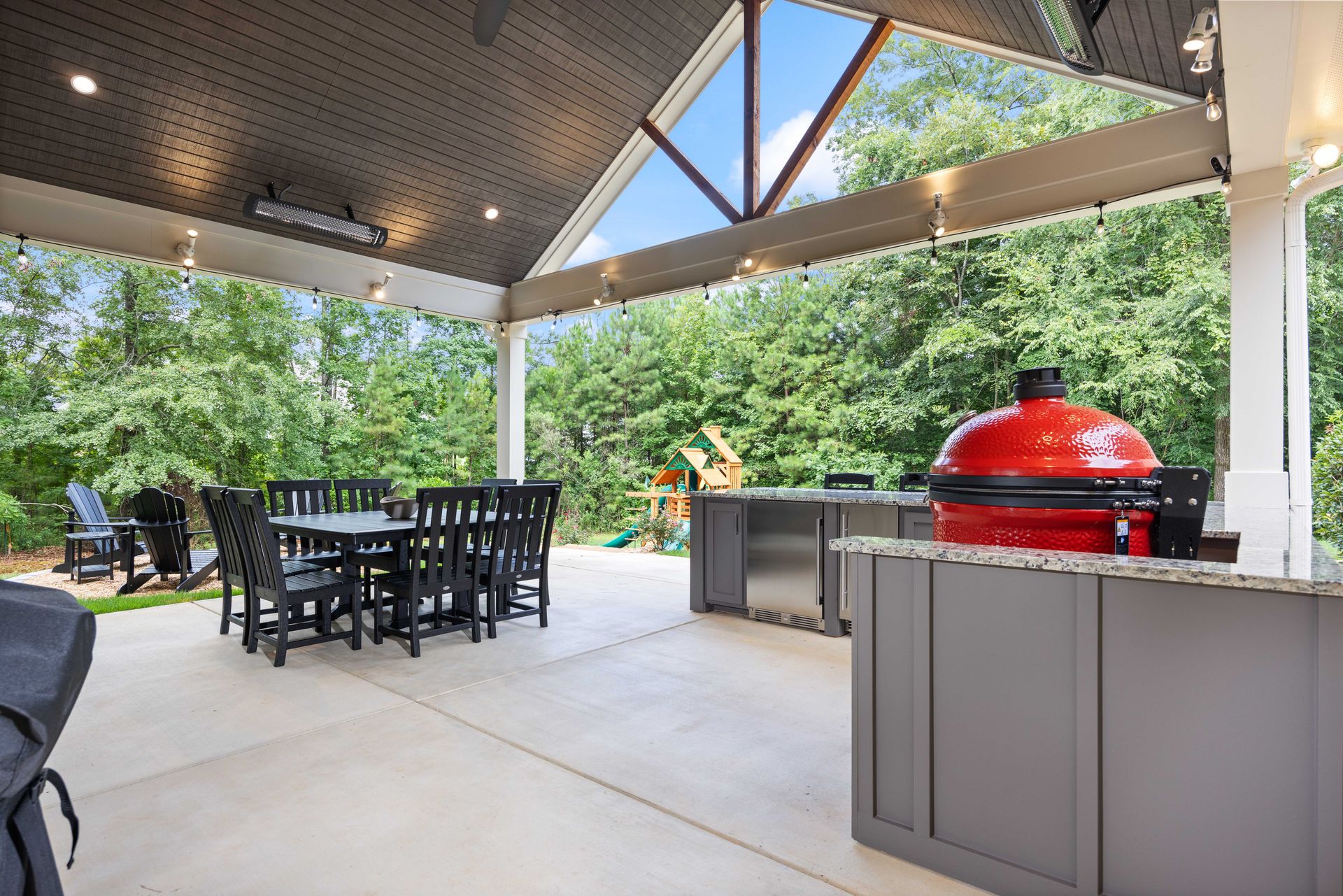 Covered patio with dining table and chairs beside a red grill, surrounded by trees