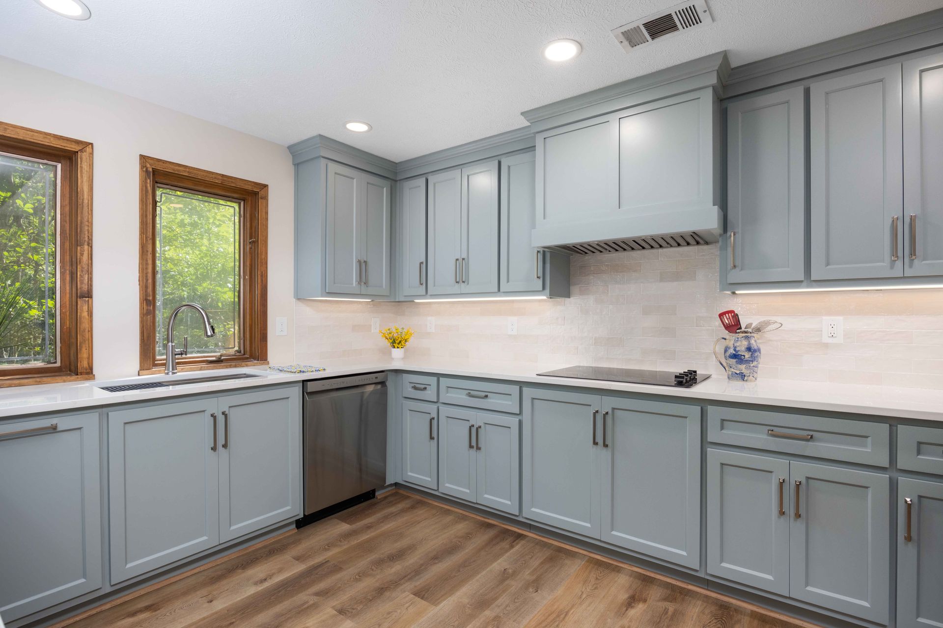 Bright modern kitchen with light blue cabinets, stainless dishwasher, wood floors, and white countertops.