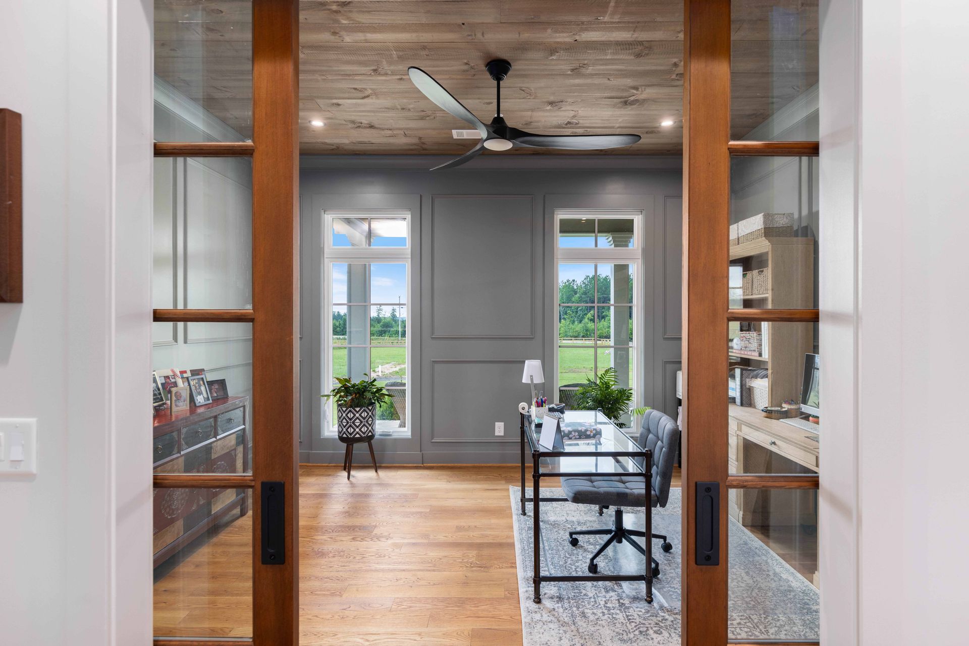 Modern home office with glass doors, wood floors, and a desk near tall windows and a ceiling fan
