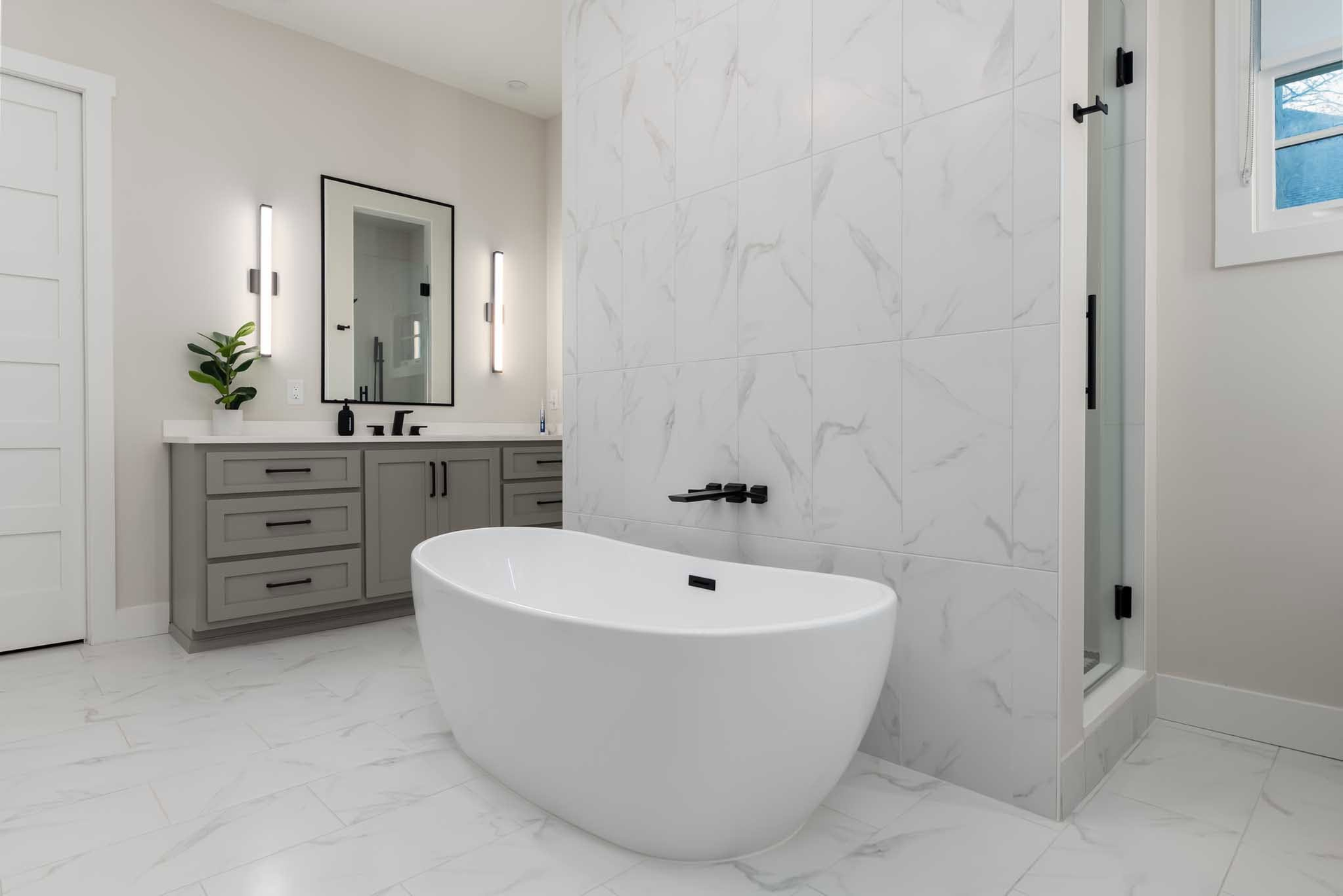 Bright modern bathroom with white marble tile, freestanding tub, gray vanity, and mirror sink area.