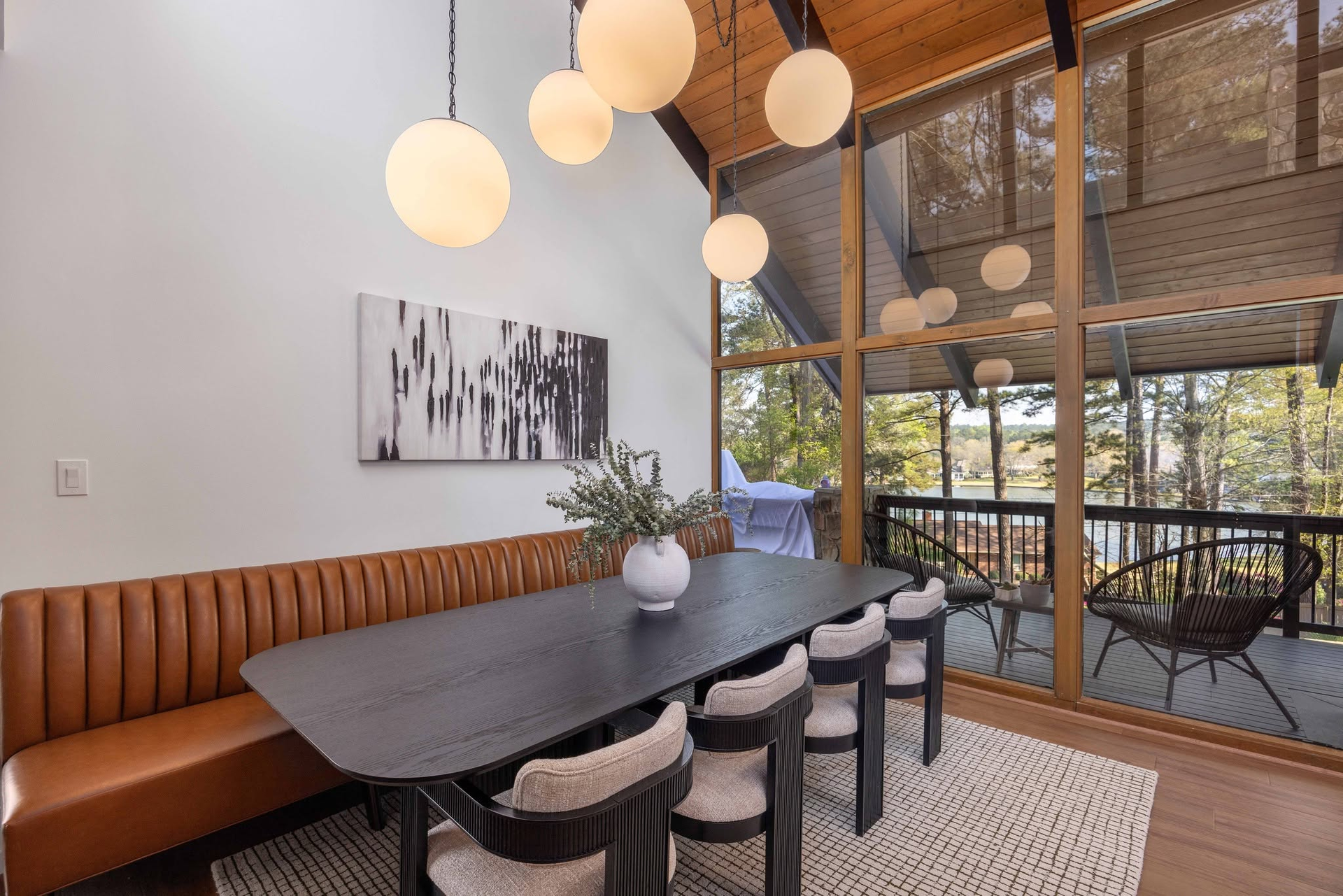 Modern dining room with a dark table, brown bench, hanging globe lights, and large windows overlooking a patio.