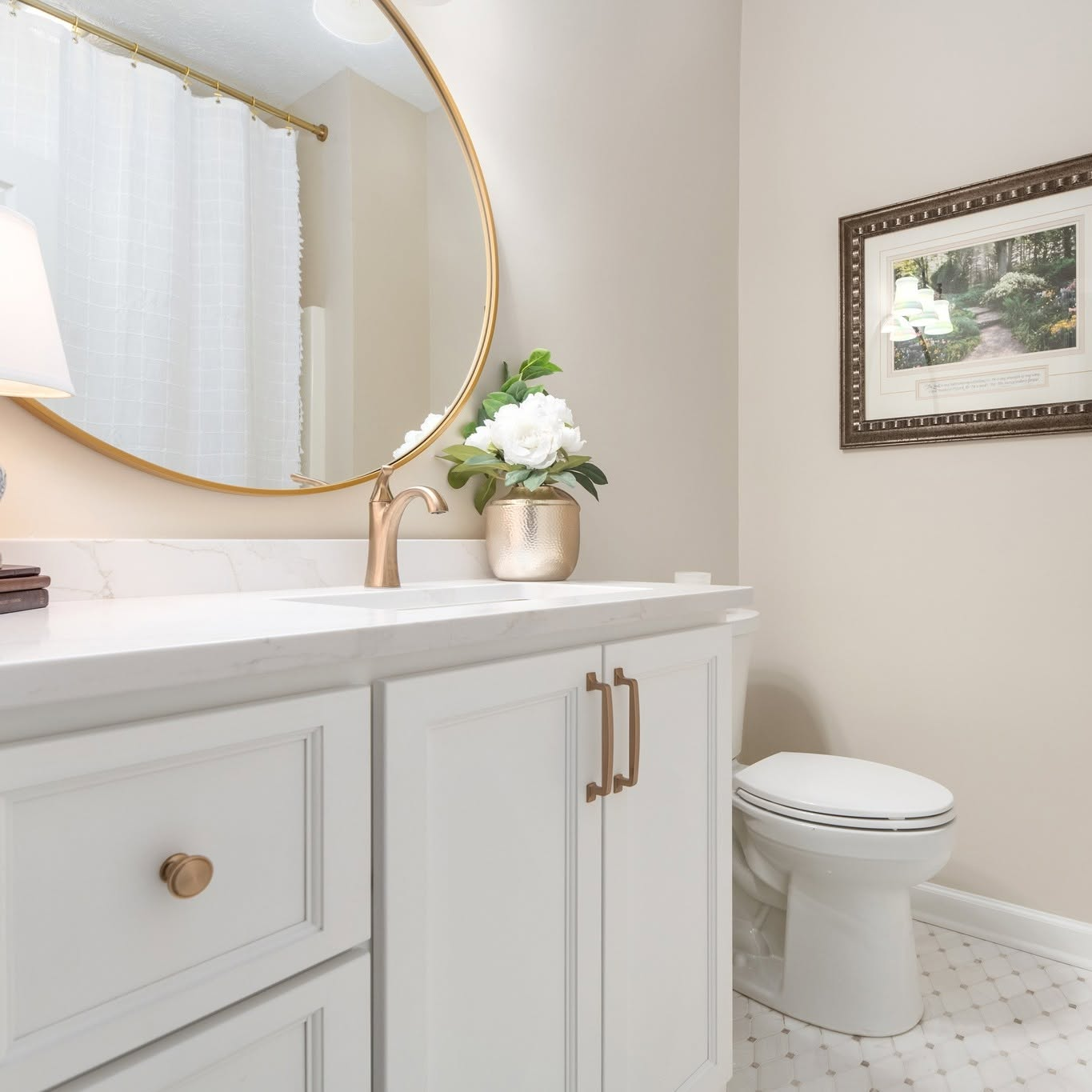 Bright, white bathroom vanity with round gold mirror, floral decor, and toilet on patterned floor.
