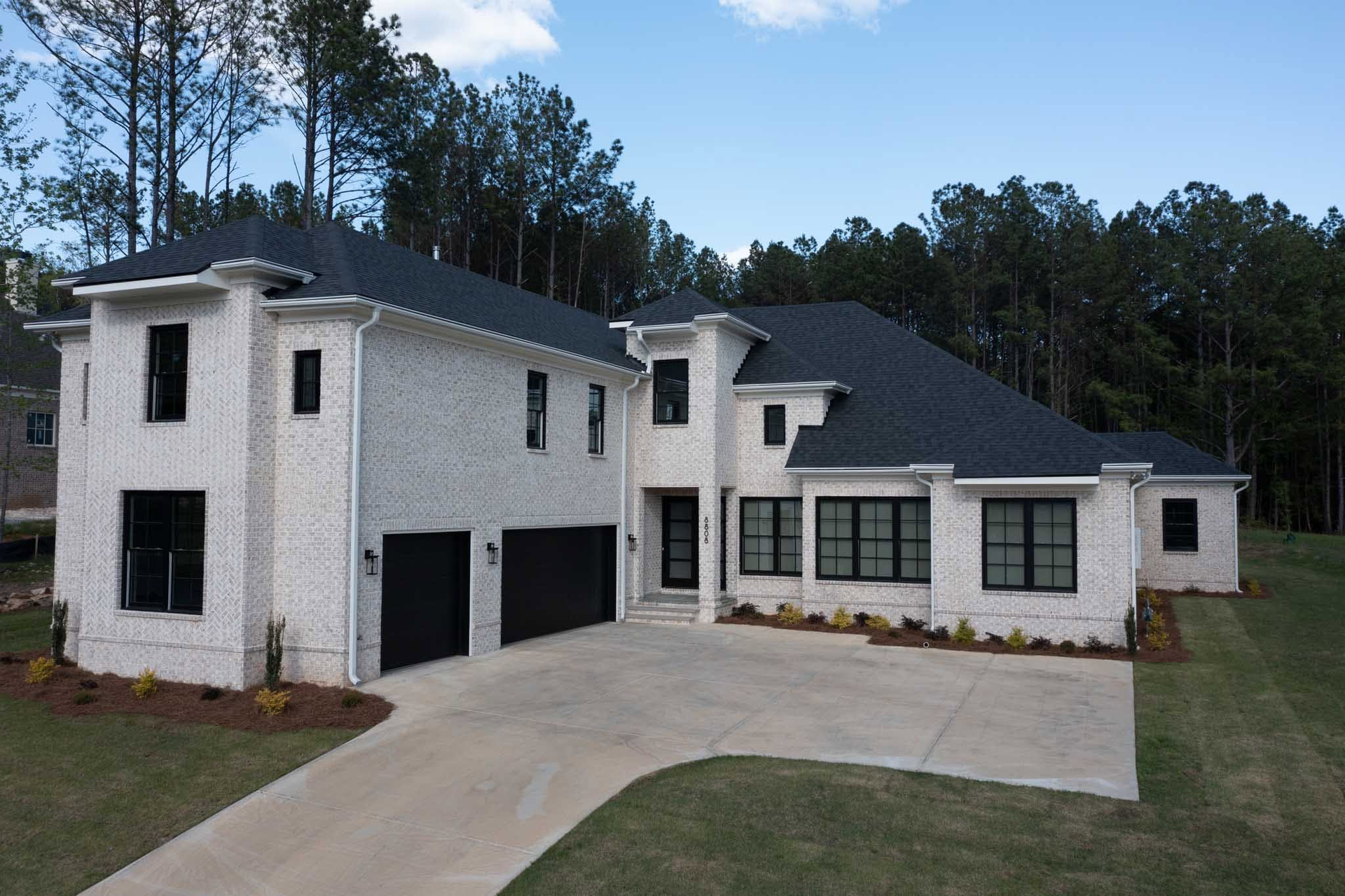 Modern two-story white brick house with black garage doors and a curved driveway under a blue sky