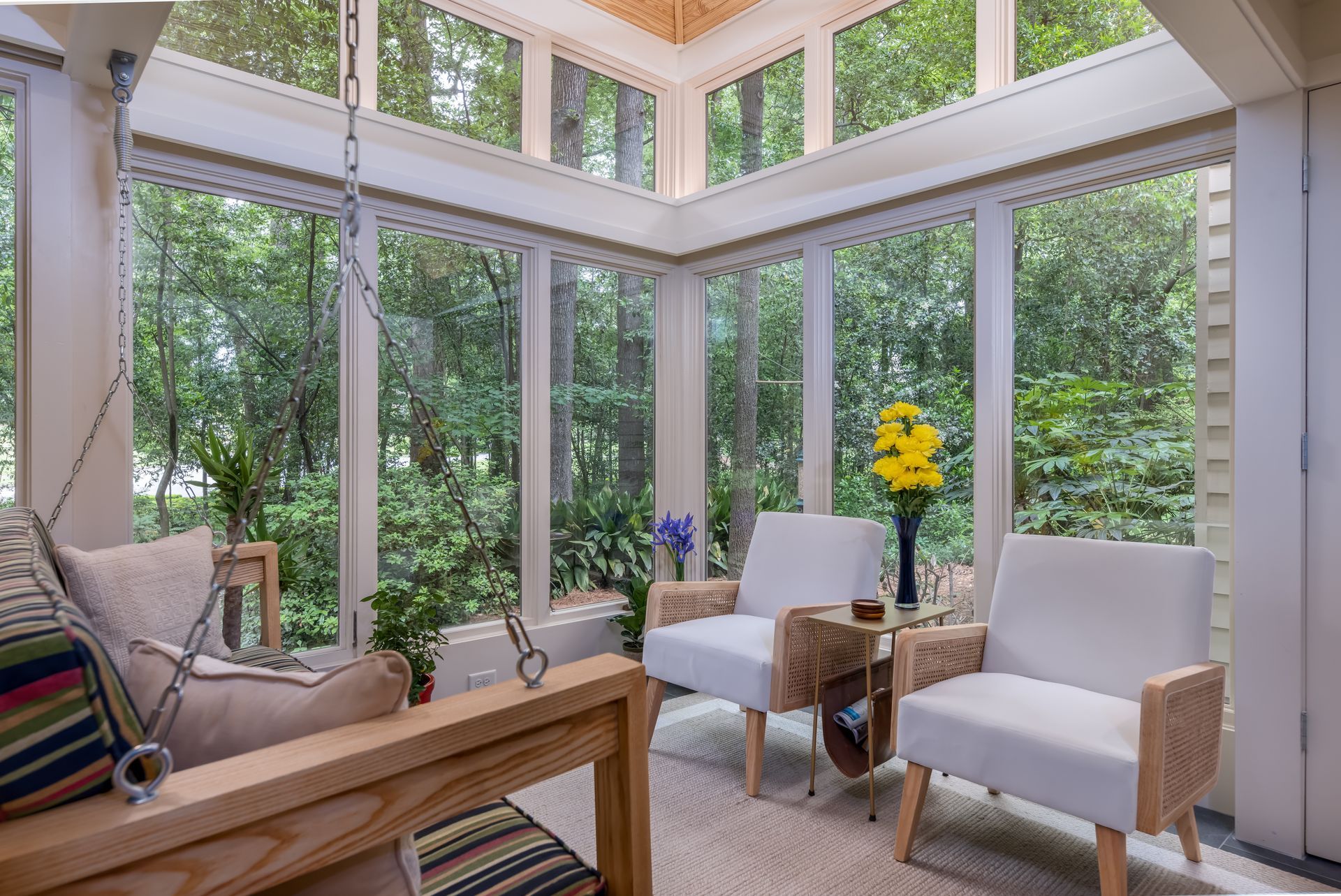 Bright sunroom with large windows, two white chairs, a wooden bench, and greenery outside.