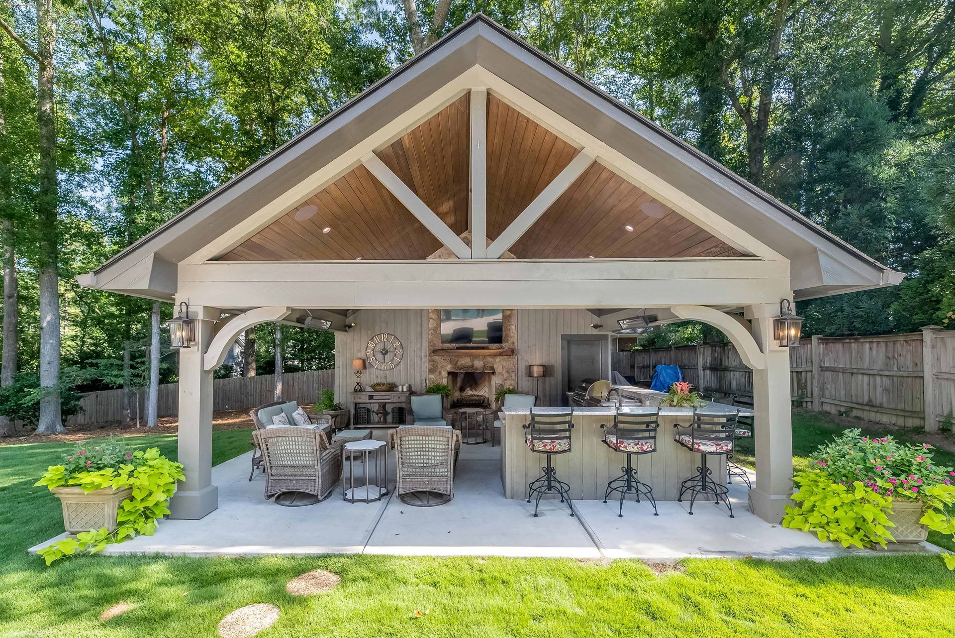 Covered outdoor patio with wicker seating and bar stools under a wooden gable roof, surrounded by greenery