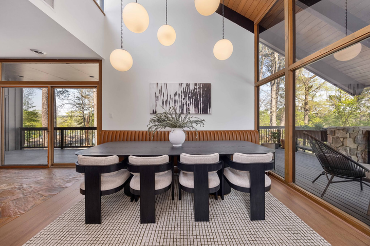 Modern dining room with a long table, black stools, hanging globe lights, and large windows overlooking trees