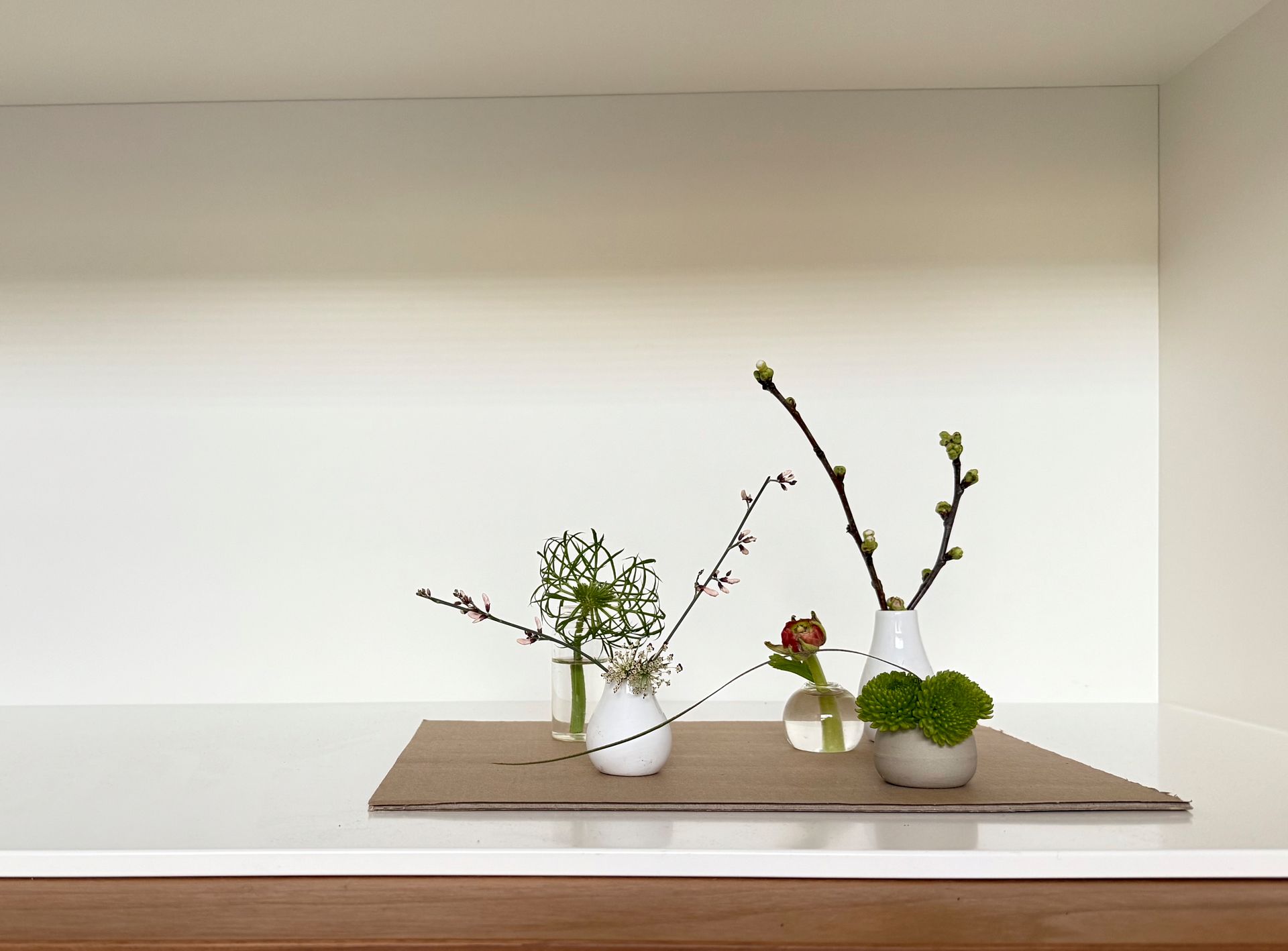 Drie kleine bloemstukjes op een neutrale achtergrond; witte vazen ​​bevatten groene takjes, knoppen en een enkele rode bloem.