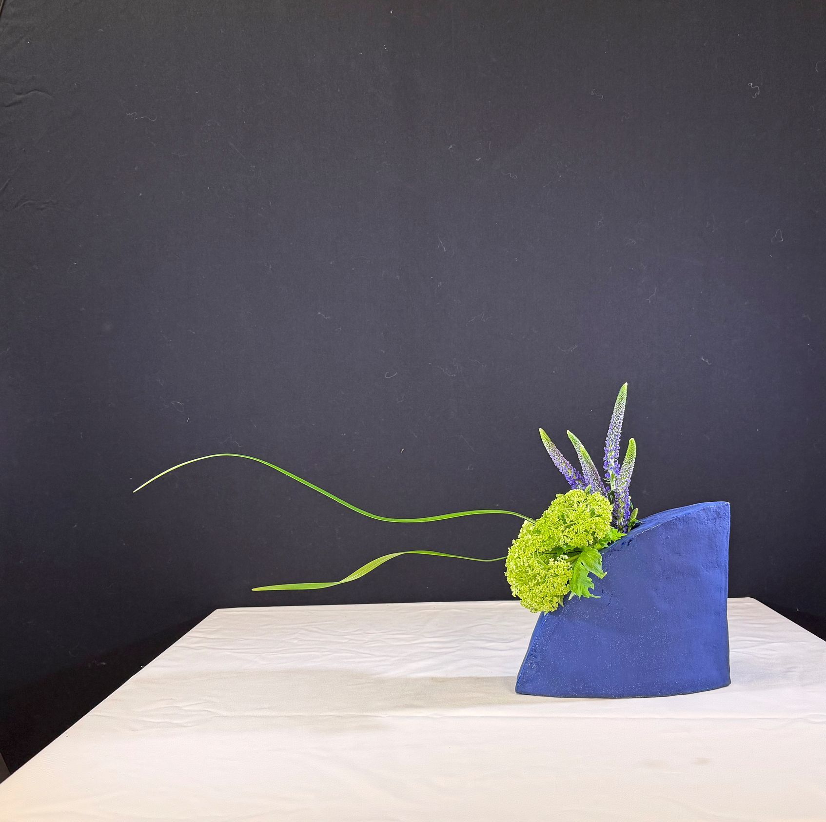 Een blauw bloemstuk op een witte tafel met een zwarte achtergrond, met groene en paarse bloemen.
